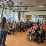 Skupina oseb v dvorani posluša nastopajočo osebo, ki stoji spredaj in vodi bralno uro.