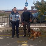 Dva policista stojita zunaj z nemškim ovčarjem, v ozadju parkirišče in modro nebo.