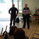 Policista predstavljata delo policije v prostoru doma, pred njima sedijo poslušalci.