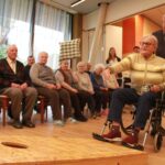 Starejša gospa v invalidskem vozičku z metom cilja v leseno tarčo z odprtino. Za njo stoji moški, ki jo spremlja. Ob strani sedi skupina starejših oseb, ki spremljajo igro.