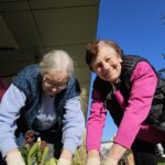 Dve osebi s rokavicami sadita rastline v cvetlično posodo in se nasmehneta v objektiv.