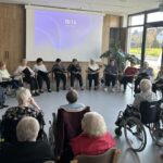 Pevska skupina nastopa v krogu, občinstvo posluša petje v skupnem prostoru doma.