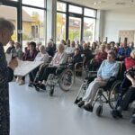 Oseba z mikrofonom vodi pevski nastop, poslušalci zbrano spremljajo izvajanje pesmi.