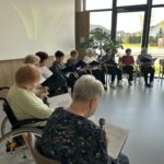 Člani pevskega zbora sedijo v krogu in pojejo ob spremljavi listov z besedili.