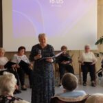 Oseba z mikrofonom poje pred skupino nastopajočih in občinstvom v dvorani doma.