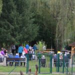 Skupina oseb se druži na prostem med drevesi, več jih sedi na klopeh in v vozičkih ob kurišču.