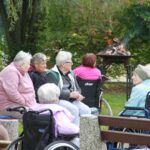 Osebe sedijo v krogu ob ognju in se sproščeno pogovarjajo na jesenskem druženju.