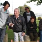 Oseba z zbranim izrazom pripravlja težko kroglo za igro, ob njej stojijo in sedijo drugi udeleženci.