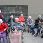 Skupina stanovalcev doma za starejše sedi zunaj pred stavbo. Nekateri so v invalidskih vozičkih, drugi na stolih. Pogovarjajo se in luščijo kostanj.