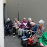 Stanovalci doma sedijo ob zidu na dvorišču doma. Klepetajo in luščijo kostanj, nekateri uporabljajo hojce in invalidske vozičke.