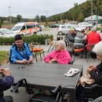 Stanovalci doma za starejše sedijo za zunanjimi mizami in luščijo kostanj. Na parkirišču v ozadju so avtomobili in reševalno vozilo.