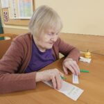 Starejša gospa v rjavi jopici sedi za mizo in z zelenim flomastrom piše na list papirja, pred njo je hruška in listi z napisom.