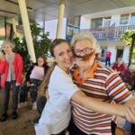 Starejša gospa in mlajša ženska skupaj plešeta in se nasmehneta v kamero. Okoli njiju stoji več ljudi, ki spremljajo dogodek.