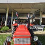 Rdeč starinski traktor parkiran pred stavbo doma. Okoli njega se zbirajo ljudje, ki sodelujejo na dogodku.