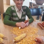 Oseba sedi za mizo in lušči koruzo, ob njej so storži in zrna.