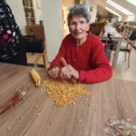 Oseba v rdečem puloverju lušči koruzo za mizo, na kateri so koruzna zrna.