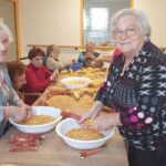 Skupina oseb sedi za mizo in skupaj lušči koruzo, smeh in prijetno vzdušje.