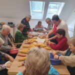 Osebe sedijo za mizo in luščijo koruzo v posode, vzdušje je delovno in veselo.