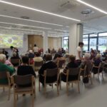 Stanovalci se zbirajo v jedilnici doma na dogodku Mini Oktoberfest, kjer je na projekciji prikazana naslovnica prireditve.