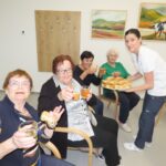 Skupina stanovalk se ob prigrizkih in pijači veselo druži z zaposleno, ki deli sladke dobrote.