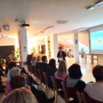 Oseba vodi predstavitev v dvorani, kjer poslušalci sledijo predstavitvi na platnu.