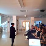 Oseba nagovarja skupino obiskovalcev, ki sedijo in poslušajo v dvorani.