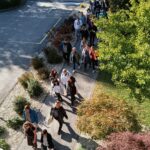 Skupina oseb hodi po pločniku ob domu v sončnem vremenu.