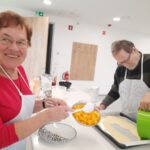 Stanovalca pri pripravi jogurtovega peciva; moški vliva maso na pekač, ženska ob njem dodaja koščke breskev in se nasmehne proti kameri.