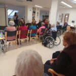 Oseba pripoveduje zgodbo občinstvu v dvorani, poslušalci so pozorni in sproščeni.