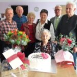 Skupina oseb stoji okoli mize z rožami in torto za praznovanje 100. rojstnega dne.