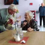 Oseba v obleki podarja šopek in darilo praznovanki, ki se nasmejano zahvaljuje.