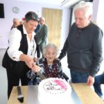 Oseba pomaga slavljenki pri rezanju torte z napisom ob 100. rojstnem dnevu.