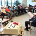 Stanovalci v dvorani spremljajo pevski nastop, v ospredju pa je rojstnodnevna torta na mizi.