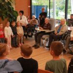 Skupina otrok stoji v vrsti in poje ali deklamira pred starejšimi stanovalci doma, ki sedijo na stolih in v invalidskih vozičkih ter jih z nasmehom spremljajo.
