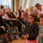 Otroci v ospredju nastopajo, stanovalci doma v ozadju sedijo v invalidskih vozičkih in spremljajo njihov nastop, nekateri se nasmihajo.