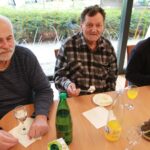 Trije starejši moški sedijo za mizo, pred njimi so krožniki s torto in kozarci pijače, eden drži žlico s kosom sladice.
