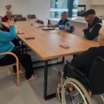 Policistka vodi pogovor s skupino starejših oseb, ki sedijo za dolgo mizo v svetlem prostoru doma starejših.
