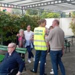 Stanovalci in zaposleni se po evakuaciji zbirajo na varnem mestu; ena od zaposlenih nosi jopič civilne zaščite.