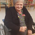 Starejša gospa v invalidskem vozičku se nasmejano ozira proti kameri in v rokah drži šopek nageljnov.