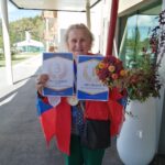 Starejša gospa v rdečem suknjiču stoji pred vhodom v dom upokojencev in se nasmejano drži dveh diplom ter šopka cvetja. Na diplomah piše »1. mesto – met na koš« in »3. mesto – met v tarčo«.