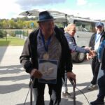 Starejši gospod s klobukom in hojco se nasmejano drži diplome za prvo mesto v šahu in nosi medaljo okoli vratu. V ozadju so drugi udeleženci športnih iger in avtomobili.
