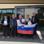 Skupina šestih starejših oseb in spremljevalke stoji pred vhodom doma upokojencev. Vsi držijo diplome in medalje, v ospredju pa je moški v vozičku z razgrnjeno slovensko zastavo.