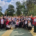 Večja skupina stanovalcev in zaposlenih doma za starejše stoji na prostem in veselo dviguje roke. Udeleženci se smejijo in držijo priznanja ter medalje, za njimi so drevesa in modro nebo.