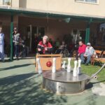 Stanovalec doma za starejše sodeluje v igri kegljanja na prostem z visečo kroglo. Okoli njega stojijo in sedijo gledalci ter zaposleni, ki spremljajo dogajanje.