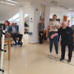 Stanovalci doma za starejše sodelujejo v igri z metanjem žogic na tarčo v skupnem prostoru. Okoli stojijo zaposleni in gledalci, ki spodbujajo igralce.