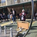 Stanovalka doma starejših drži rdečo kroglo in se pripravlja na met kegljev na zunanjem igrišču doma za starejše. Okoli nje sedijo in stojijo drugi stanovalci ter zaposleni.