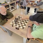 Dva moška sedita za mizo in igrata šah v skupnem prostoru doma starejših. Eden kaže na šahovnico, drugi ga opazuje. V ozadju sedijo še drugi igralci.