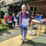 Nasmejana udeleženka stoji na dvorišču doma, v roki drži priznanje in nosi medaljo za prvo mesto v ruskem kegljanju.