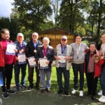 Skupina stanovalcev in zaposlenih stoji na prostem, vsi nasmejani držijo priznanja in medalje, za njimi je lesena uta okrašena s koruzo.