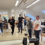 Skupina stanovalcev in zaposlenih stoji v prostoru, eden od moških izvaja met žogice proti tarči, drugi ga opazujejo in spodbujajo.